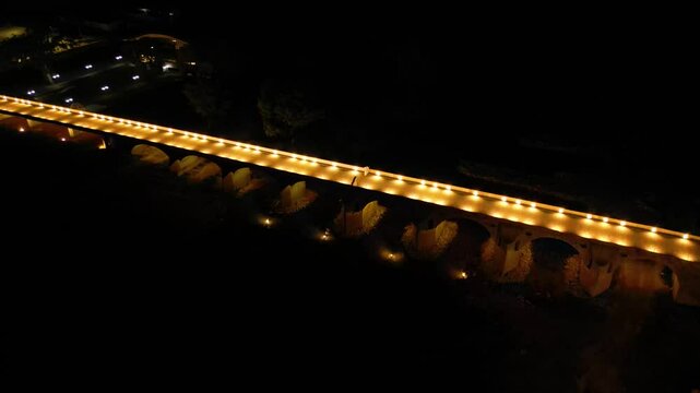 Aerial high angle footage of a 150 years old stone masonry bridge illuminated on a dark night. Drone shot of Master Kolyo Fitcheto's bridge at Byala village over the river of Yantra in Bulgaria.
