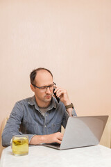 A man sits at a table, intently typing on his laptop while a glass of drink sits nearby. He is...