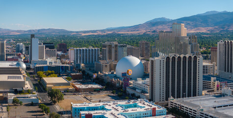 Daytime aerial view of downtown Reno, Nevada, showcasing the silver dome of the National Bowling Stadium, high rise buildings, and Sierra Nevada mountains. © Aerial Film Studio