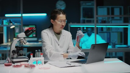 Asian female scientist in lab coat, safety goggles and gloves examining red liquid in test tube and typing on laptop while conducting tissue engineering research in biotech laboratory