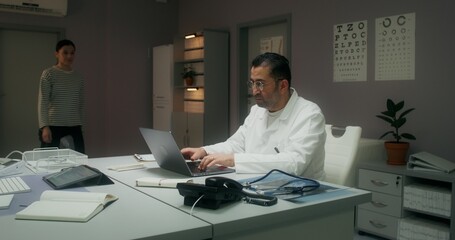 A female patient enters a medical office for a medical appointment. The doctor uses a laptop sitting at the table