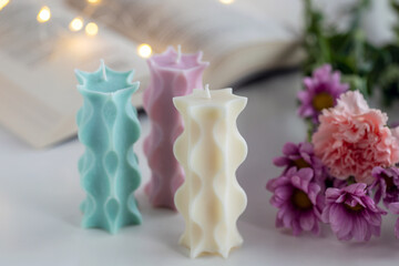 Three soy candles on a table with a book, flowers and lights in background. Home decor.