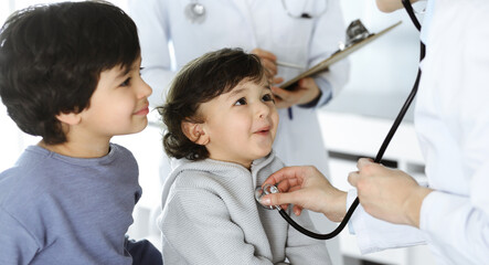 Woman-doctor examining a child patient by stethoscope. Cute arab toddler and his brother at physician appointment. Medicine concept