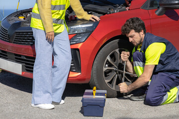 Young professional mechanic changing flat tire broken vehicle on the road with reflective clothing,...