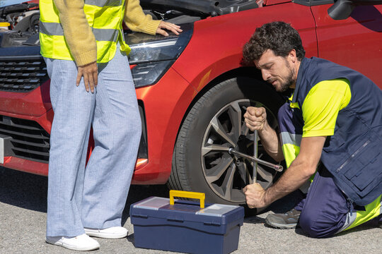 Young professional mechanic changing flat tire broken vehicle on the road with reflective clothing, toolbox and cross wrench, broken engine, roadside assistance, insurance tow truck
