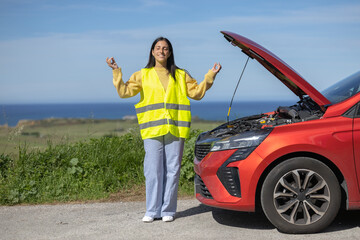 Pretty young woman in happy reflective vest feels at peace with broken car on the road while...