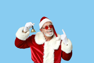 Santa Claus in sunglasses with Christmas bell pointing at something on blue background