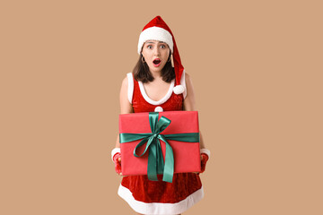 Shocked young woman in Santa Claus costume with Christmas gift box on brown background