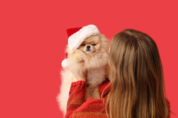 Young woman with cute Pomeranian dog in Santa hat on red background