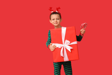 Cute little boy in pajamas with candy canes and Christmas gift on red background