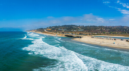 Aerial view of San Diego's coastline featuring a sandy beach, a prominent bridge, and a developed area with buildings. Vehicles and people are visible.