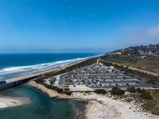 Aerial view of San Diego's coastal area featuring a beach, parking lot, road, bridge, ocean waves, and hillside residential buildings under a clear sky.