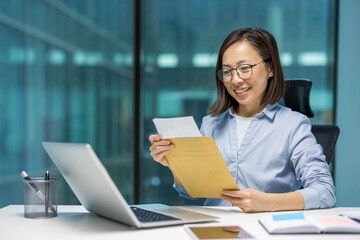 Asian businesswoman reads good news from envelope at office desk, surrounded by laptop and documents, showcasing professional success and positivity