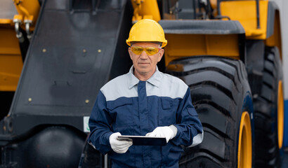 Industrial engineer uses computer tablet to manage production. Adult man in uniform and yellow helmet against background of mining tractor © Parilov
