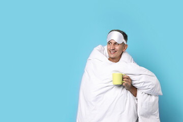 Happy man with sleep mask and cup wrapped in blanket on light blue background, space for text