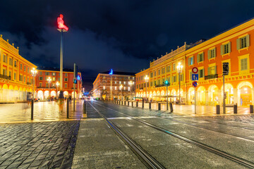 The illuminated Place Massena square with the seven color statues on poles and distant city in view behind the train tracks in Nice, France, on the French Riviera Cote d'Azur.