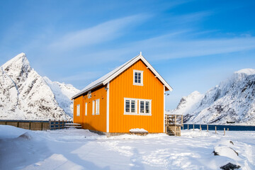 Traditional yellow rorbu house and Olstind mountain peak in Sakrisoy fishing village on Lofoten islands, Norway. Winter landscape with snowy mountains and Scandinavian cottage