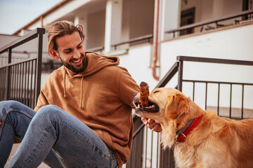 young attractive and sporty guy in casual clothes, sitting outside in home garden playing and having fun with his pet dog during a spring day at sunset	