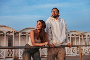 a couple of engaged people dressed in casual clothes are leaning on the handrail of a beach establishment during a spring afternoon at sunset