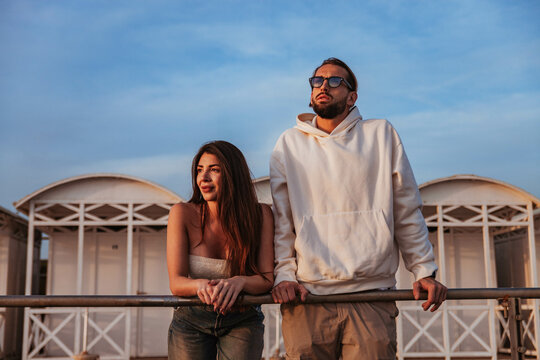 a couple of engaged people dressed in casual clothes are leaning on the handrail of a beach establishment during a spring afternoon at sunset
