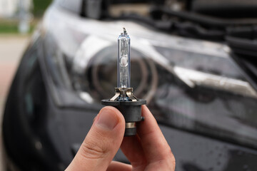A man's hand holds a single xenon bulb against the blurred background of a car headlight. Replacing the bulb yourself is an important part of ensuring road safety and visibility at night.