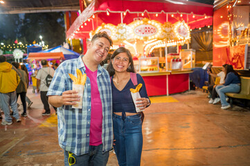 Couple of Latino friends posing in front of the camera during their visit to the amusement park, in their hands they hold food made with artisanal ingredients.