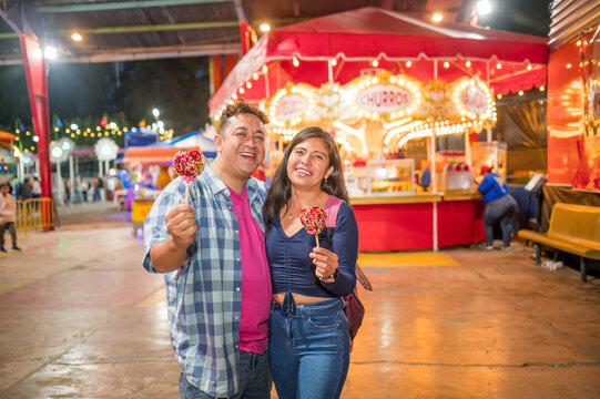 Couple of Latino friends having fun at their local fair, smiling at the camera with food in their hands that they have purchased from local vendors.
