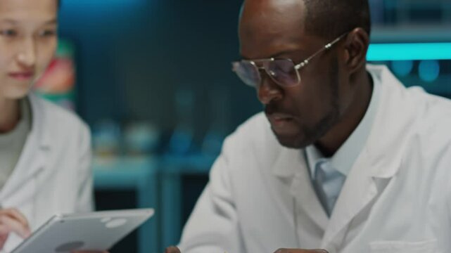 Tilt up shot of Black food scientist eating burger made with cultured meat and describing its taste to Asian female colleague as she using tablet during laboratory test