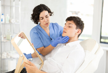 Fototapeta premium Young male patient lying on clinical chair discussing face aesthetic procedures with doctor
