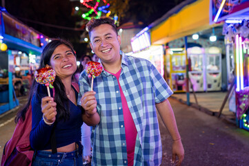 A couple smiles at the camera, holding a red apple with caramel in their hands. They have visited the fair in their city during the evening.