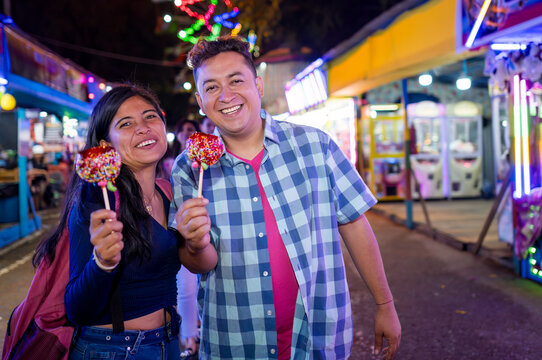 A couple smiles at the camera, holding a red apple with caramel in their hands. They have visited the fair in their city during the evening. - Powered by Adobe
