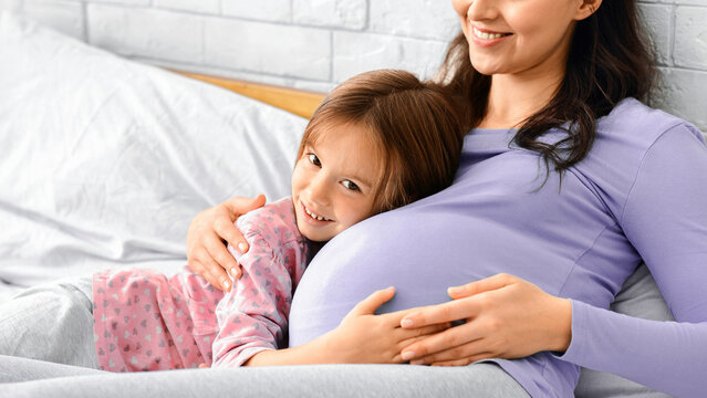 Little daughter and pergnant mother in pajamas bonding on bed, girl hugging mommy tummy, copy space