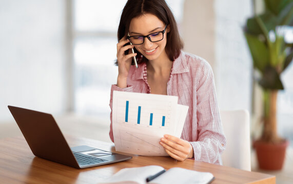 Business Lifestyle. Young female manager consulting client on phone, holding papers, checking financial report in office