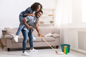 Joyful Spring-Cleaning. Playful African American Couple Fooling Together During Tidying Flat, Woman Piggybacking Her Boyfriend While He Mopping Floor