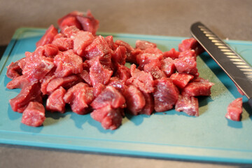 Close up of raw beef meat chopped in small cubes on green cutting board with a knife