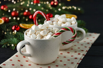 Tasty hot cocoa drinks with candy canes and marshmallows in cups on black wooden table, closeup