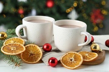Tasty hot cocoa drinks in cups, fir branches, baubles and dry orange slices on white table against blurred lights, closeup