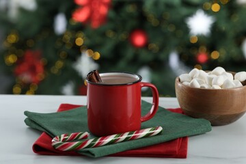 Tasty hot cocoa drink with cinnamon sticks in mug, candy cane and marshmallows on white table against blurred lights