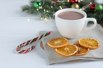 Yummy cocoa in cup, candy cane, dried orange slices and Christmas wreath on white wooden table. Space for text