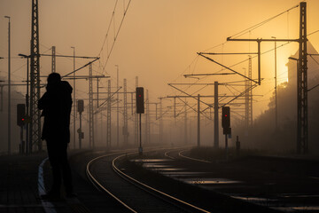 The train platform glows softly in the morning light, with dawn's golden hue wrapped in mist. A red train slowly arrives, ready for the early crowd.
