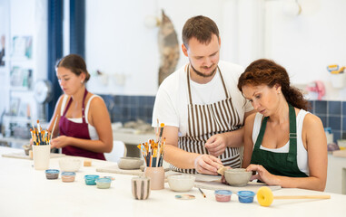 In pottery workshop, apprentice female students sitting at table, engaged in making products from...