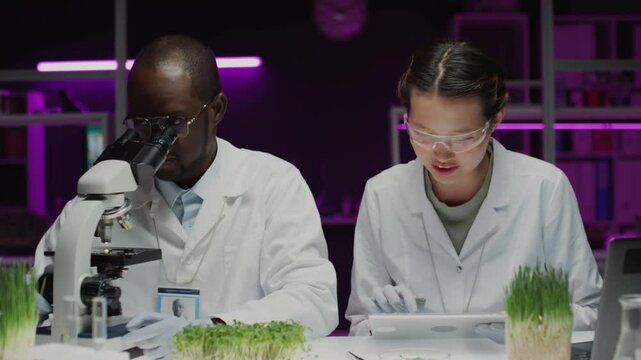 African American male botanist talking with Asian female colleague, then looking through microscope as she using digital tablet during plant genetics research in laboratory