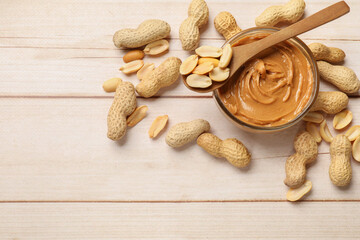 Tasty peanut butter in bowl and spoon with groundnuts on wooden table, flat lay. Space for text