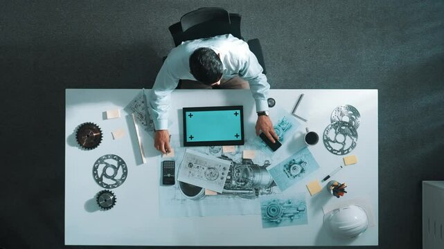 Top view of engineer looking at phone while working on tablet with green screen at meeting table with turbine engine or jet engine sketch design and various metal gear scatter around. Alimentation.