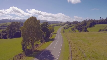 A winding paved road leads through a lush green valley with a blue sky and white clouds overhead.