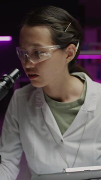 Vertical shot of Asian female scientist in gloves and goggles looking through microscope and taking notes on clipboard during research in laboratory
