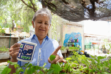 A woman holds a cup and touches some plants. She smiles and seems to be amused.