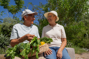 A couple of older people are sitting on a bench and holding a basket of vegetables. They are smiling and seem to be enjoying their time together
