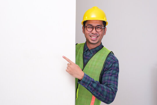 A construction worker peeking behind wall pointing beside him with happy expression