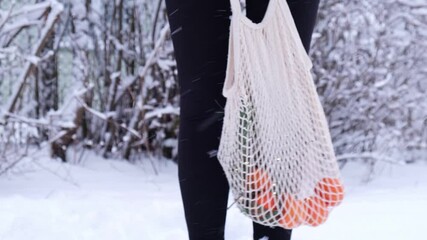 Unrecognizable woman holding Mesh shopping bag with tangerines in snowy winter forest outdoor. Go green concept eco-friendly sustainable Christmas. Responsible celebration 
